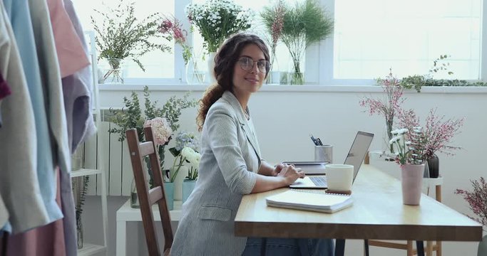 Smiling young business woman professional fashion designer, florist, decorator using laptop at workplace with flowers. Female wedding planner looking at camera working in creative studio. Portrait.