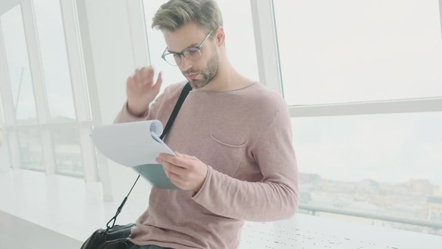 A concentrated young bearded man wearing glasses is reading the documents while standing near the window inside