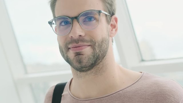 A handsome pleased young bearded man wearing glasses is looking to the side while standing near the window inside
