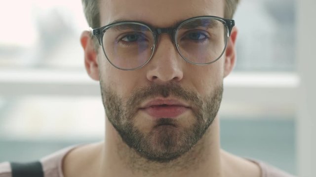A calm young bearded man wearing glasses is opening his eyes while standing near the window inside