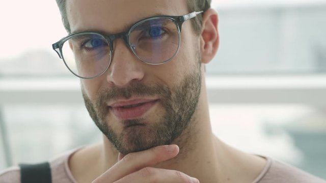 A handsome young bearded man wearing glasses is posing to the camera while standing near the window inside
