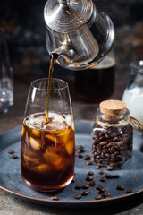 Ice coffee cold summer drink in a tall glass with pouring coffee and coffee beans on a stone background.