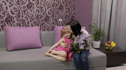 Little daughter and mother sitting on sofe with bouquet of lilac purple flowers