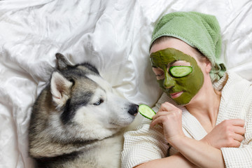 a woman with a towel on her head lies on a bed with a dog. on her face a green clay cosmetic mask with cucumbers. funny home spa with a pet. wellness concept © Maryna