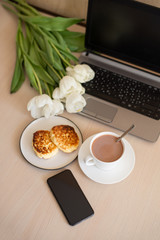 view above phone, laptop, Cup with cacao and saucer on the table, white tulips, homemade cheesecakes on a plate  (vertically)