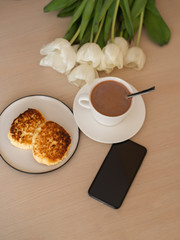 above phone, laptop, Cup with cacao and saucer on the table, white tulips, homemade cheesecakes on a plate