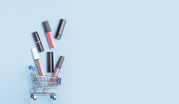 Women's lipsticks and lip glosses in a shopping trolley on a blue background. The concept of buying cosmetics, online shopping.