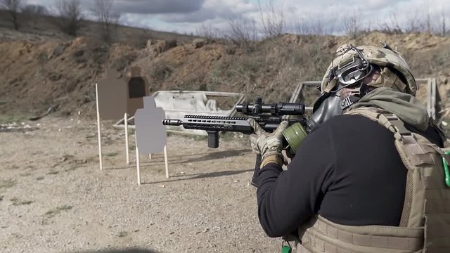 Unknown soldier in a gas mask, helmet and life vest shoots from an assault rifle with a scope. A rookie trains at the training range outdoor. A supervisor watches him. Bullets fly out from the gun.