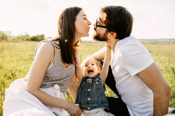 Family of three, mother, father and little daughter, sitting on a blanket in a meadow. Little girl trying to stop parents from kissing.
