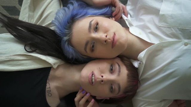 Beautiful lesbian women posing for photo shoot lying on floor in modern interior. Spbd Portrait of young homosexual couple is looking at camera with smile and touching faces with hands, showing