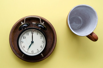 cup for tea or coffee and a black analog alarm clock on a brown ceramic plate on a yellow background. The concept of toning in the morning.