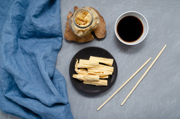 space for text. fermented soy tofu Yuba skin with spices on a brown plate and in a jar on a gray neutral background with bamboo sticks, soy sauce and a towel. Asian cuisine