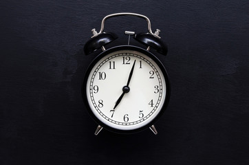 a black alarm clock with a white dial shows the time of 7 hours on a black slate background with place for text