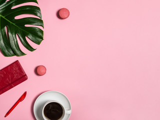 Female workspace with red notebook and pen, coffee cup, macarons and green monstera leaf on pink background, top view