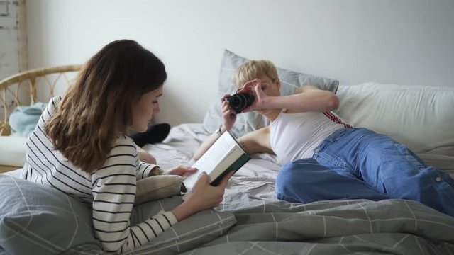 Young lesbian girl photographs her girlfriend lying on bed in home room. Spbd Short-haired woman is making photo holding camera in hand, female is reading book and posing with smile in light interior