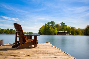 Two Muskoka chairs sitting on a wood dock facing a lake. Across the calm water is a brown cottage...