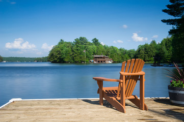 Muskoka chair sitting on a wood dock facing a calm lake. Across the water is a cottage nestled among green trees. 