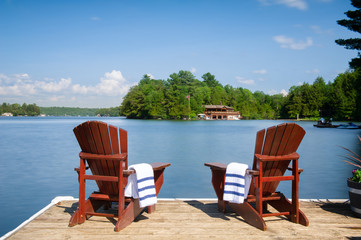 Two Muskoka chairs sitting on a wood dock facing a calm lake. Across the water is a cottage nestled among green trees. On the chairs there are towels.