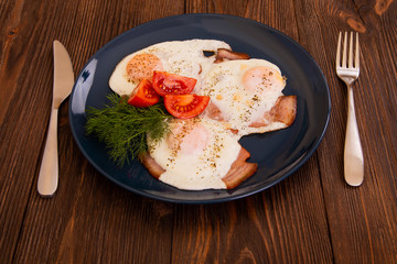 Delicious spicy snack from fried eggs, pork ham, cut tomatoes and dill on the wooden background with fork and knife