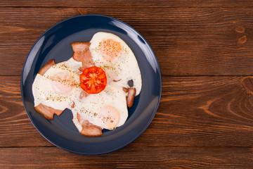 Tasty spicy breakfast with fried eggs, bacon and cut tomato in a dark plate on the wooden table, top view