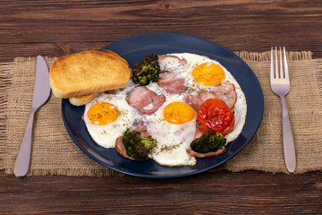 Savoury breakfast with fried eggs, bacon, tomato, broccoli and toasts in a dark plate that is on textured burlap, with fork and knife, angle view