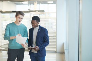 Fototapeta premium Waist up portrait of two young businessmen discussing work documents while standing in while office interior, copy space