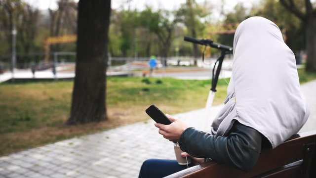 Back view of young moderm attractive Muslim woman sitting in park wearing hijab and listening to music with smartphone and cup of coffee in her hands, slow motion