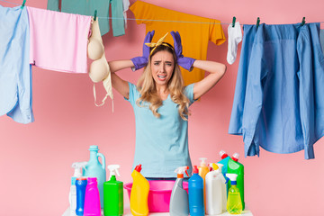 Sad housewife holds head with hands, in front of her is basin and cleaning supplies, behind is clean clothes on rope