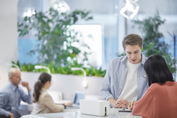 Waist up portrait of young man talking to Asian friend while standing at tall desk, copy space