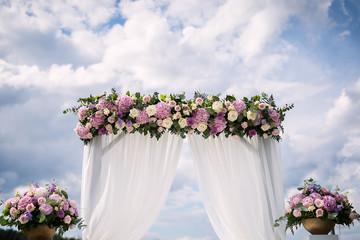 against the blue sky stands a wedding arch decorated with flowers, two vases with flowers stand on white pedestals, festive decor on a vadba, wedding of the newlyweds