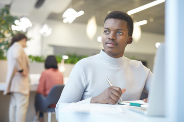 Portrait of smiling African-American man looking away dreamily while working with laptop in office,...