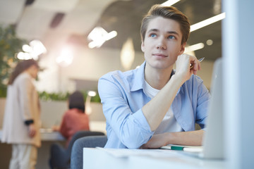 Portrait of smiling young man looking away dreamily while working with laptop in office, copy space