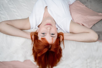 Obraz na płótnie Canvas to stay at home, not to go out, a girl with red hair is lying on the bed, tipping her head, holding her hands behind her head, looking forward, bored during self-isolation,