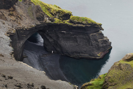 Nordic landscape. Black sand volcanic beach, rocky cape, green hill and stone hole cave on coast. Aerial view. Vestmannaeyjar island in Iceland.