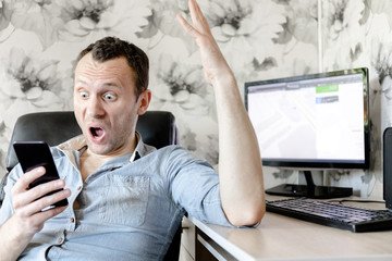 young man sits at a computer table and looks in amazement at the smartphone