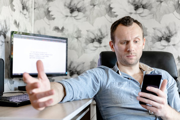 young male freelancer at home at a computer reads news on a smartphone with a puzzled look