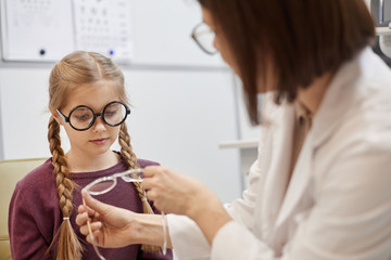 Portrait of cute teenage girl trying on glasses during vision test in modern ophthalmology clinic,...