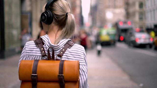 Slow motion of young woman dressed in casual wear listening music from playlist in headphones while walking on street of modern city.Back view of hipster gir strolling in urban setting 
