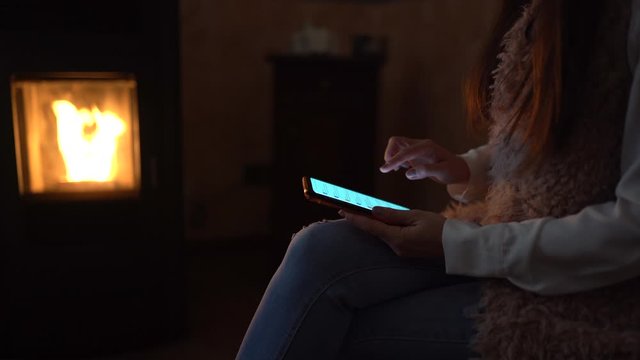 Active girl using modern cellphone and doing shopping online with modern apps sitting on the chair at home on background of burning fire in pellet stove. Modern online technologies, distance working