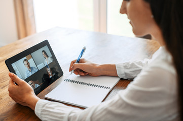 Webinars, online conference, video meeting. Young woman using app on tablet for video connect with a many people at same time together at office, she is doing notes in notebook. Distant work concept