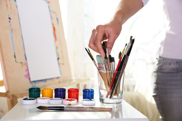 the artist’s hand takes a brush from a glass against the background of an easel