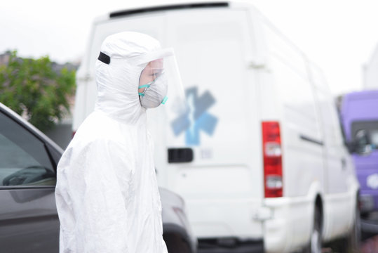 Doctor On Call. Ambulance Driver.Tired Doctor Ambulance In A Protective Antivirus Suit, A Face And Eye Mask Half A Turn To The Camera, In The Background An An Emergency Car.Help For Patients