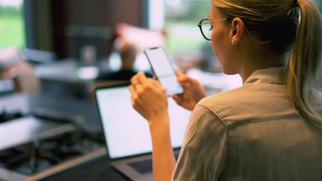 Young woman in eyeglasses typing text message on smartphone and chatting in social networks using 4G internet.Hipster girl blogging on cellular sitting at  laptop computer
