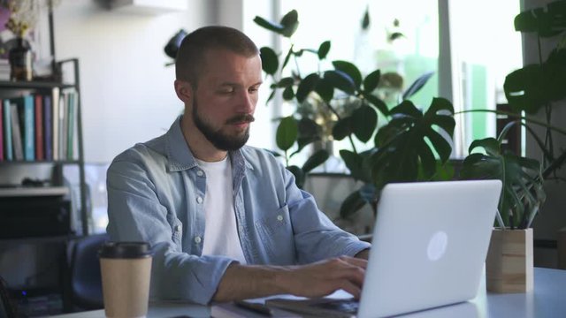 Serious businessperson handsome guy is typing using Spbd laptop computer indoors in office room busy with accounting job. paper coffee cup. man worker and freelancer student