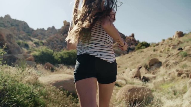 Beautiful young woman with long brown hair in modern clothing runs in slow motion through a rocky valley in the afternoon.
