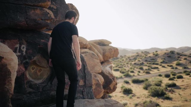 Handsome man with a beard and black clothes walks onto a monzogranite rock formation in the desert and looks to the horizon.