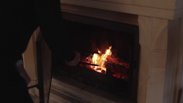 Old man taking care of the burning fire inside fireplace with metal plyers indoors