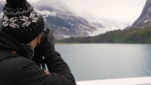 A young male photographer wearing a black coat and bobble hat stood onboard ship looking across the ocean at snow capped mountains and glaciers taking photographs on his canon camera.
