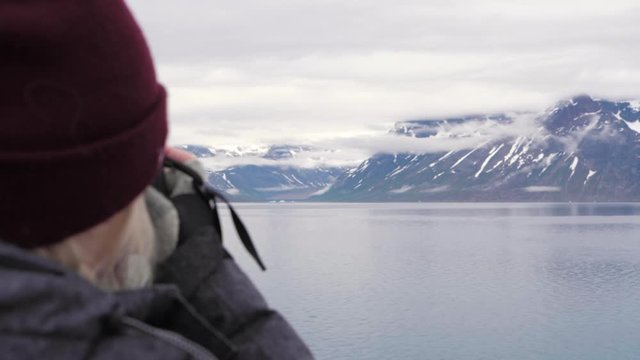 A young female photographer wearing a grey coat and bobble hat stood onboard ship looking across the ocean at snow capped mountains and glaciers taking photographs on her canon camera in slow motion