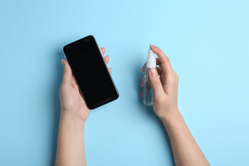 Woman spraying antiseptic onto smartphone on light blue background, top view